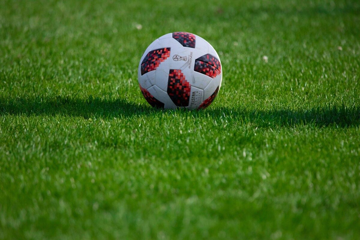 Ein weißer Fußball mit roten und schwarzen Mustern liegt auf einem grünen Rasen.