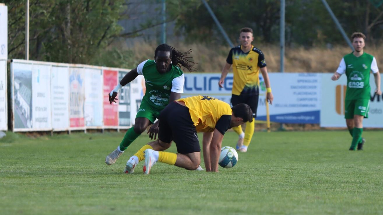 Ein Fußballspieler in einem grünen Trikot läuft auf den Ball zu, während ein anderer Spieler in einem gelben Trikot versucht, ihn zu blockieren. Ein dritter Spieler in einem gelben Trikot beobachtet von hinten.