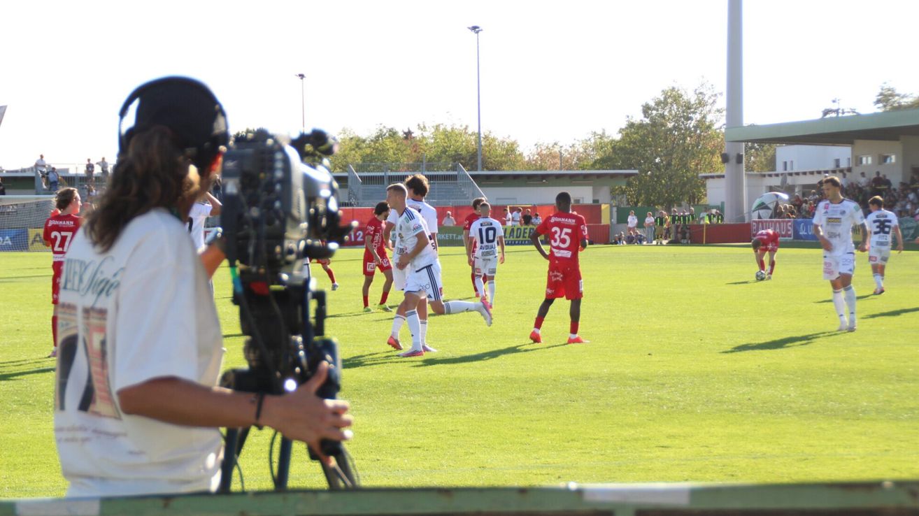 Fußballspieler in roten und weißen Trikots laufen auf dem Feld. Ein Kameramann filmt die Aktion.