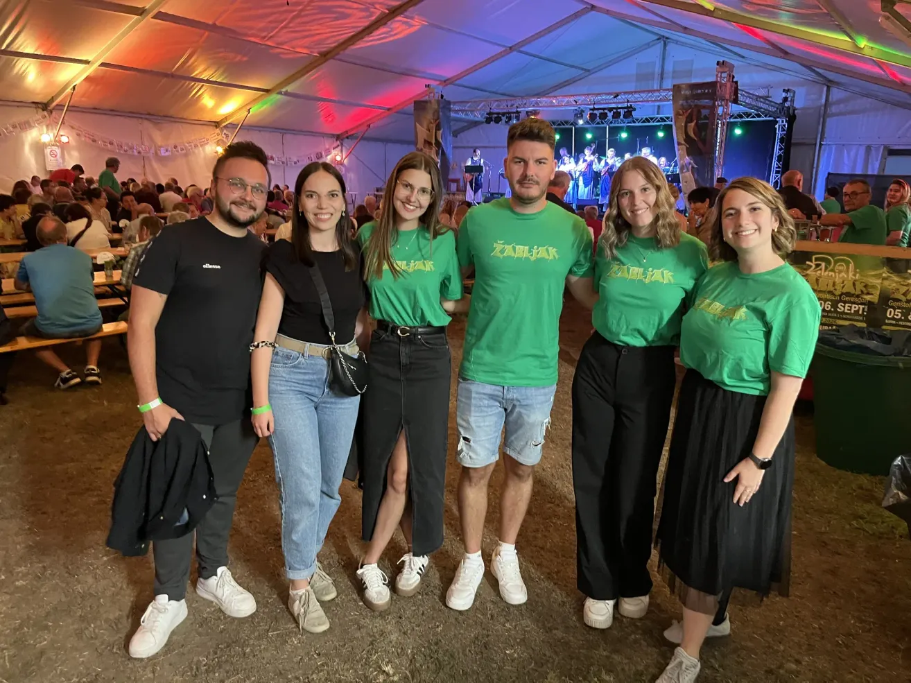 Five people in matching green shirts with the word Zabijak on them stand together in front of a stage with colorful lights.