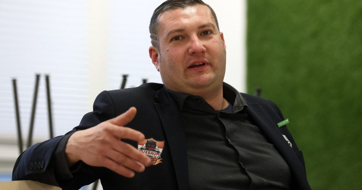 Ein Mann in einem schwarzen Jackett mit einem Logo auf der linken Brust gestikuliert, während er spricht.
