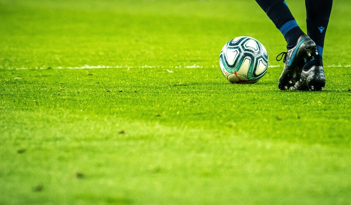 Ein Fußball liegt auf einem grünen Rasen, mit einem Fuß und Schuh des Spielers im Hintergrund.