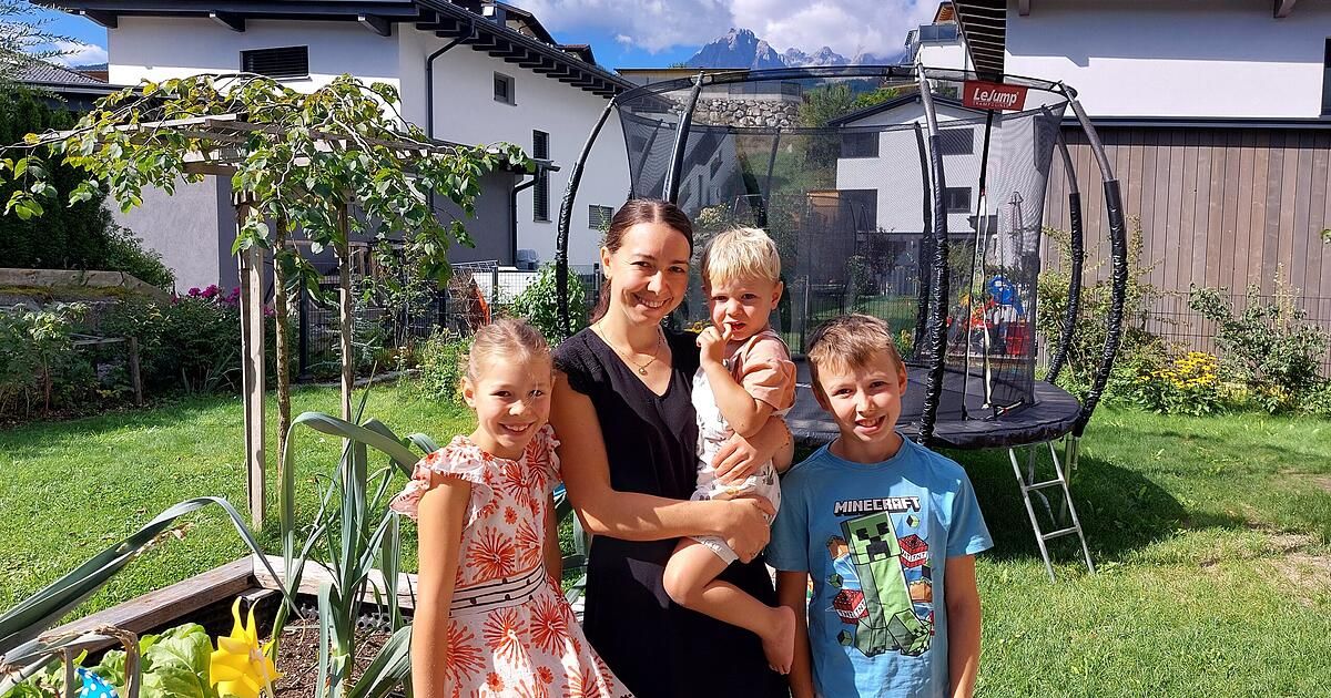 A woman and her three children stand in a garden with a trampoline in the background. The woman is smiling and holding a baby. The children are standing beside her, smiling.