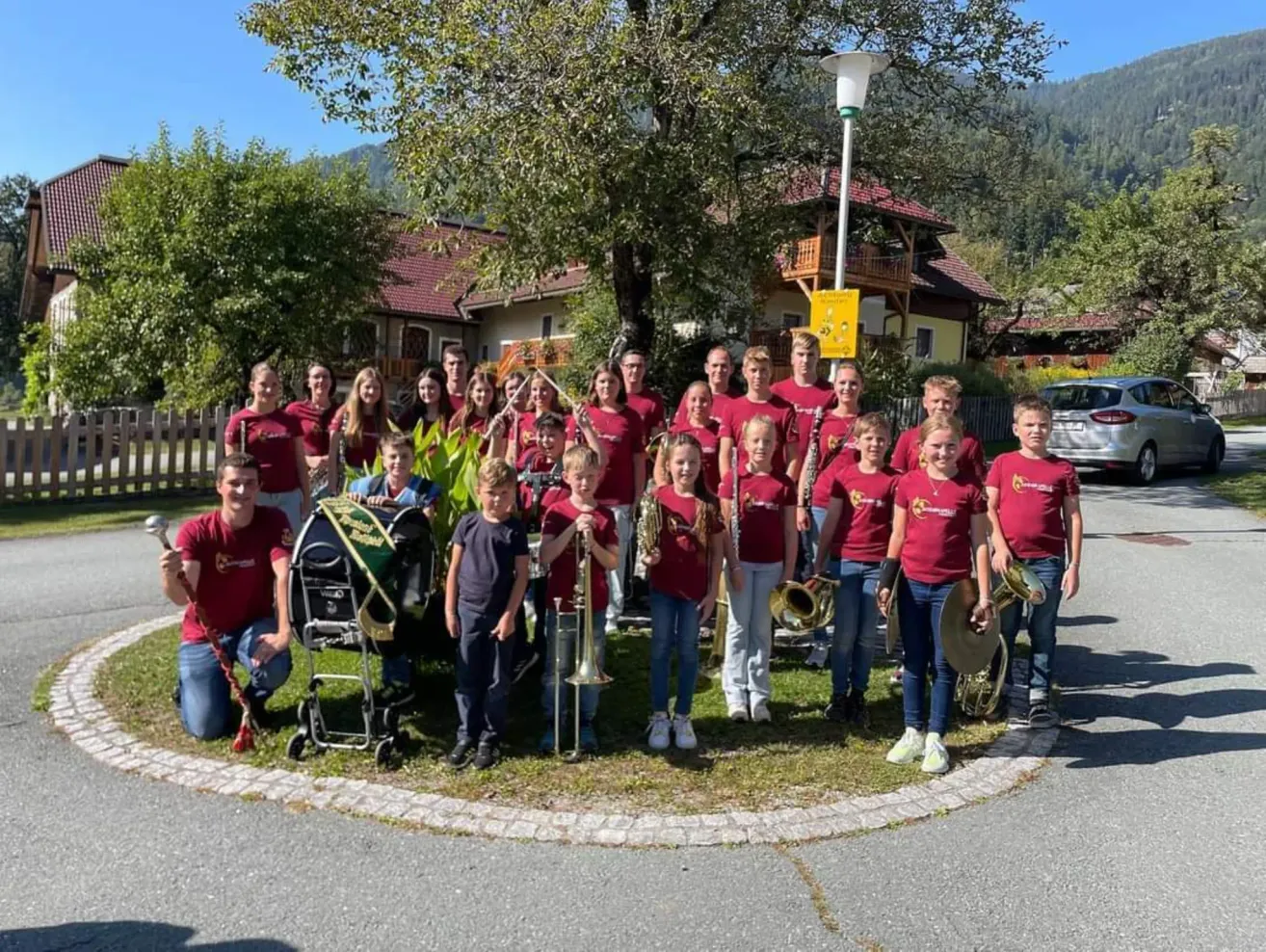 Eine Gruppe junger Musiker, gekleidet in roten T-Shirts, posiert für ein Foto auf einem Rasenstück. Sie halten verschiedene Musikinstrumente, darunter Trompeten, Posaunen und Trommeln. Hinter ihnen steht ein Haus mit einem roten Dach und einem gelben Schild.