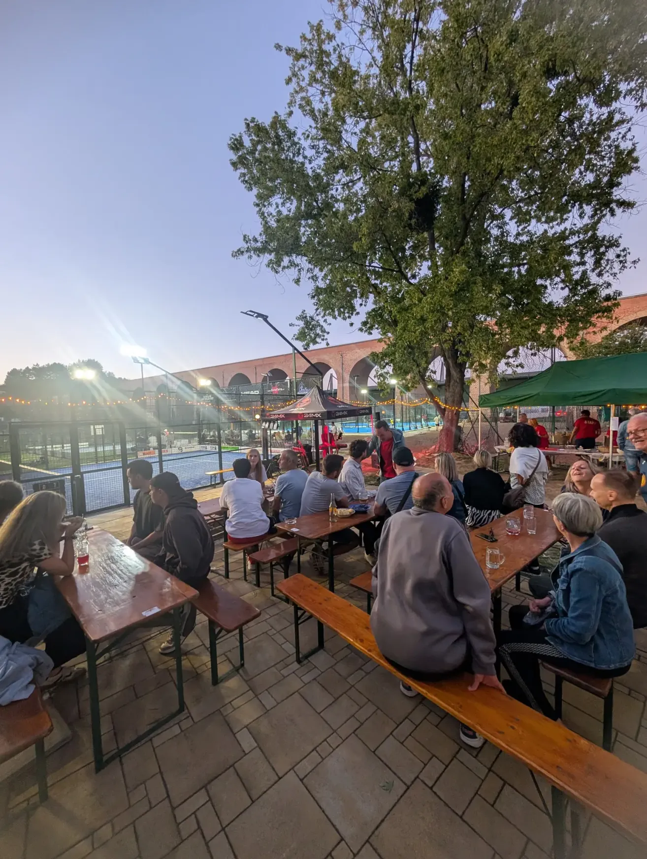 A group of people sit around wooden tables outside under a large tree, with a tennis court and bridge in the background.
