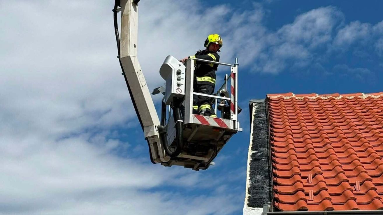 A firefighter stands on a ladder attached to a crane, wearing a yellow helmet and a reflective safety suit, working on a roof.