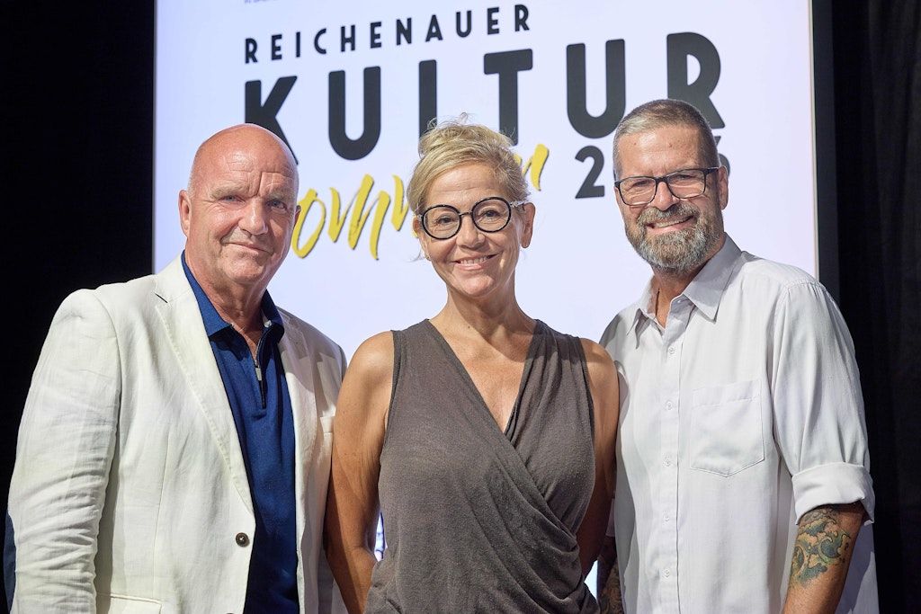 Three individuals stand together in front of a sign for 'Reichenauer Kultur'. The man on the left wears a white blazer, the woman in the middle has glasses and a sleeveless top, and the man on the right wears a white shirt. They all appear to be smiling.