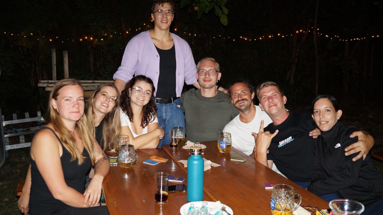 A group of people are sitting around a table at night, smiling and posing for a photo. They have drinks in front of them, and one man is making a peace sign.