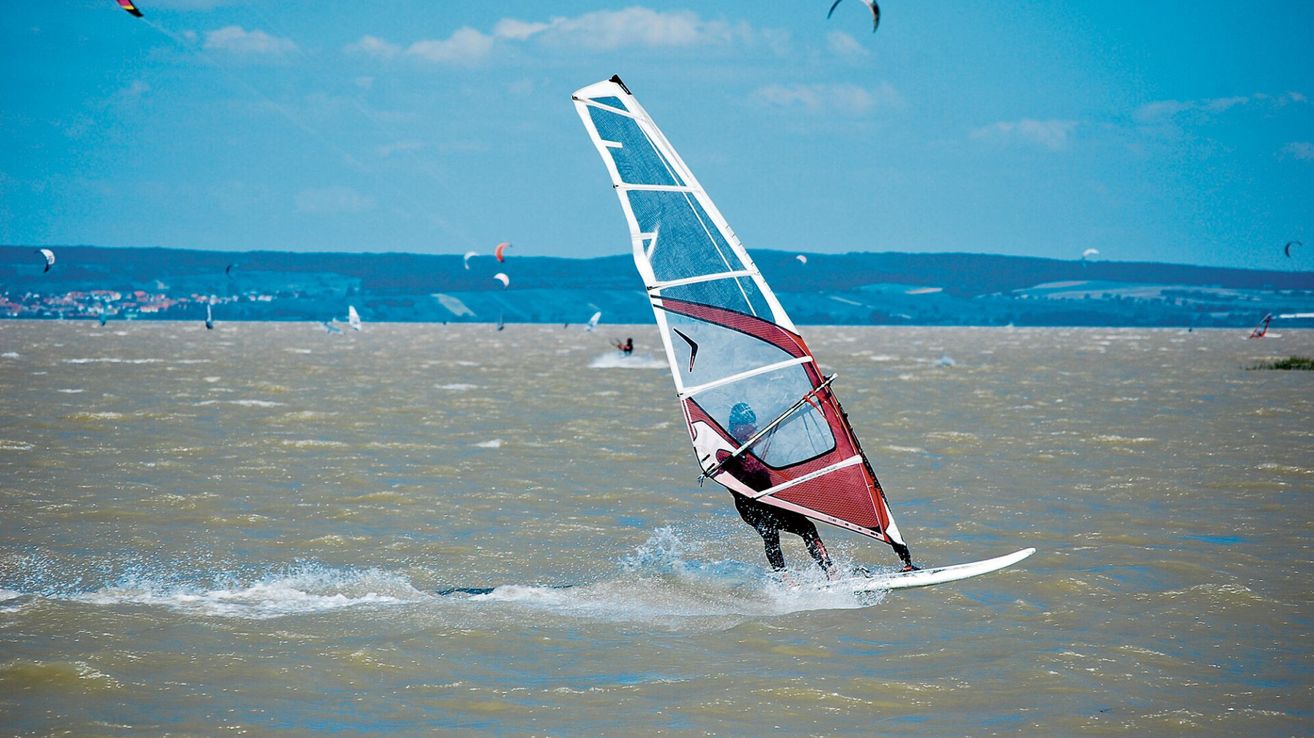Eine Person windsurft auf einer ruhigen Wasserfläche mit einem blau-roten Segel. In der Ferne sind mehrere andere Windsurfer und Berge zu sehen.