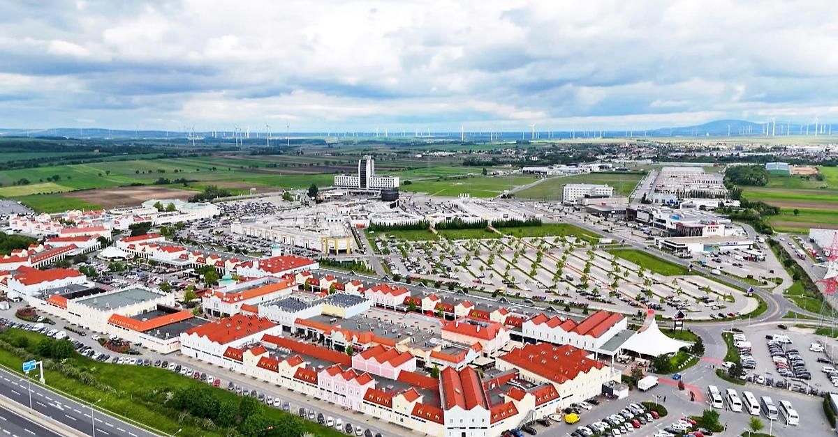 Ein Luftbild eines modernen Einkaufskomplexes mit roten Dächern, umgeben von Parkplätzen und Grünflächen, unter einem bewölkten Himmel.