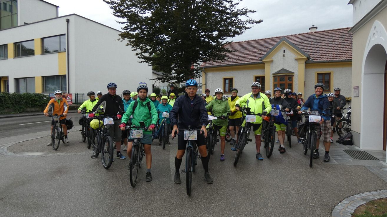 Eine Gruppe von Radfahrern, alle mit Helmen und Fahrradausrüstung, stehen in einer Reihe vor einem Gebäude. Sie scheinen an einer Radsportveranstaltung teilzunehmen.