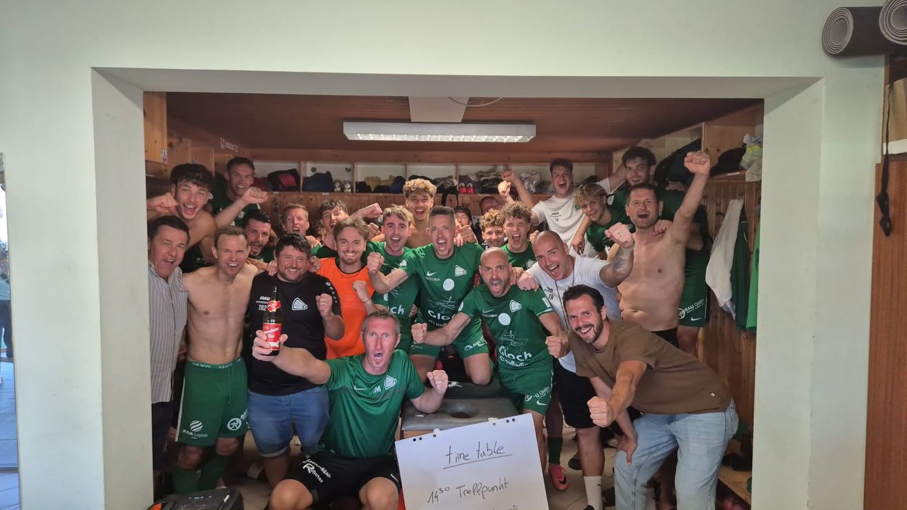 Eine Gruppe von Fußballspielern in grünen Uniformen feiert in einer Umkleidekabine, einige ohne Hemd, mit einem Mann, der eine Flasche in der Hand hält. Ein Whiteboard mit 'Zeitplan' und '1430 Treffpunkt' ist sichtbar.