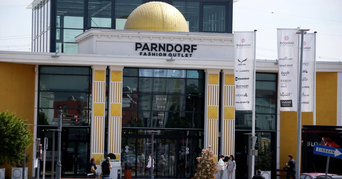 Das Äußere des Parndorf Fashion Outlets mit einer goldenen Kuppel, gelben und weißen Säulen und einer Glasfassade. Menschen gehen draußen herum.