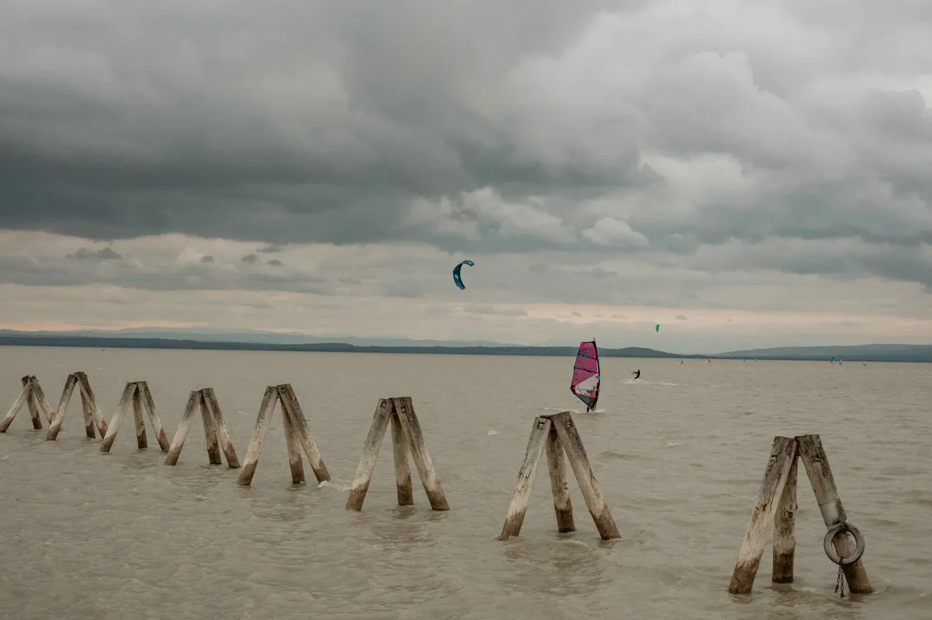 Eine Gruppe von Windsurfern navigiert durch ein ruhiges Meer mit einem bewölkten Himmel darüber. Holzpfähle säumen den Wasserrand.