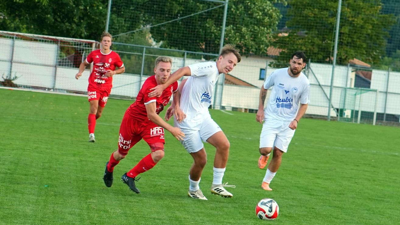 Vier Fußballspieler sind auf einem Feld, zwei in roten und zwei in weißen Trikots. Der Spieler in Rot läuft auf den Ball zu, während der Spieler in Weiß versucht, ihn zu blockieren.