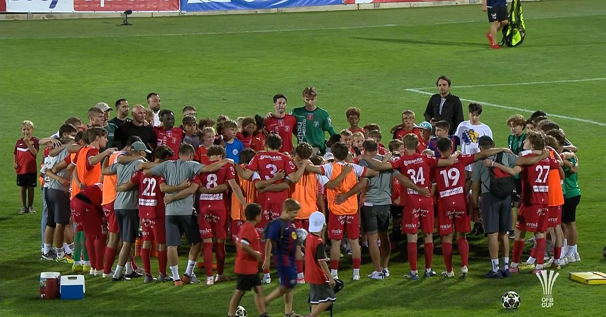 Eine Fußballmannschaft in roten und orangefarbenen Trikots ist auf einem Feld versammelt, mit Trainern und Spielern um sie herum. Die Spieler tragen Nummern auf dem Rücken.