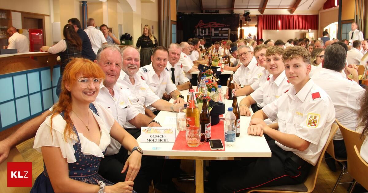 Eine Gruppe von Menschen in Uniform sitzt an einem langen Tisch, mit Flaschen, Gläsern und einem Handy auf dem Tisch. Sie lächeln und scheinen bei einer formellen Veranstaltung zu sein.