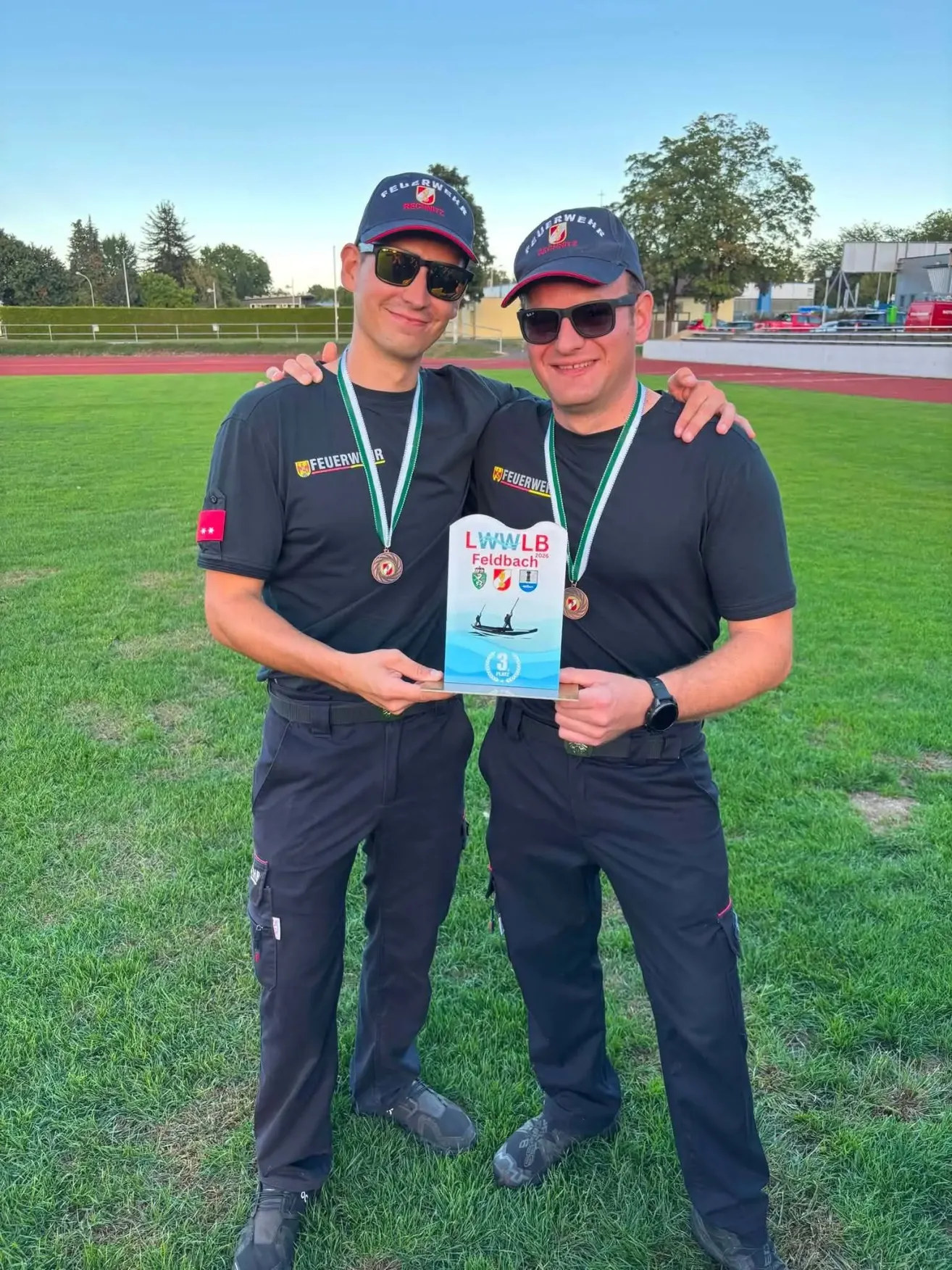 Two men wearing uniforms and hats stand on a field, holding a plaque and medals. They are smiling and posing for a photo.