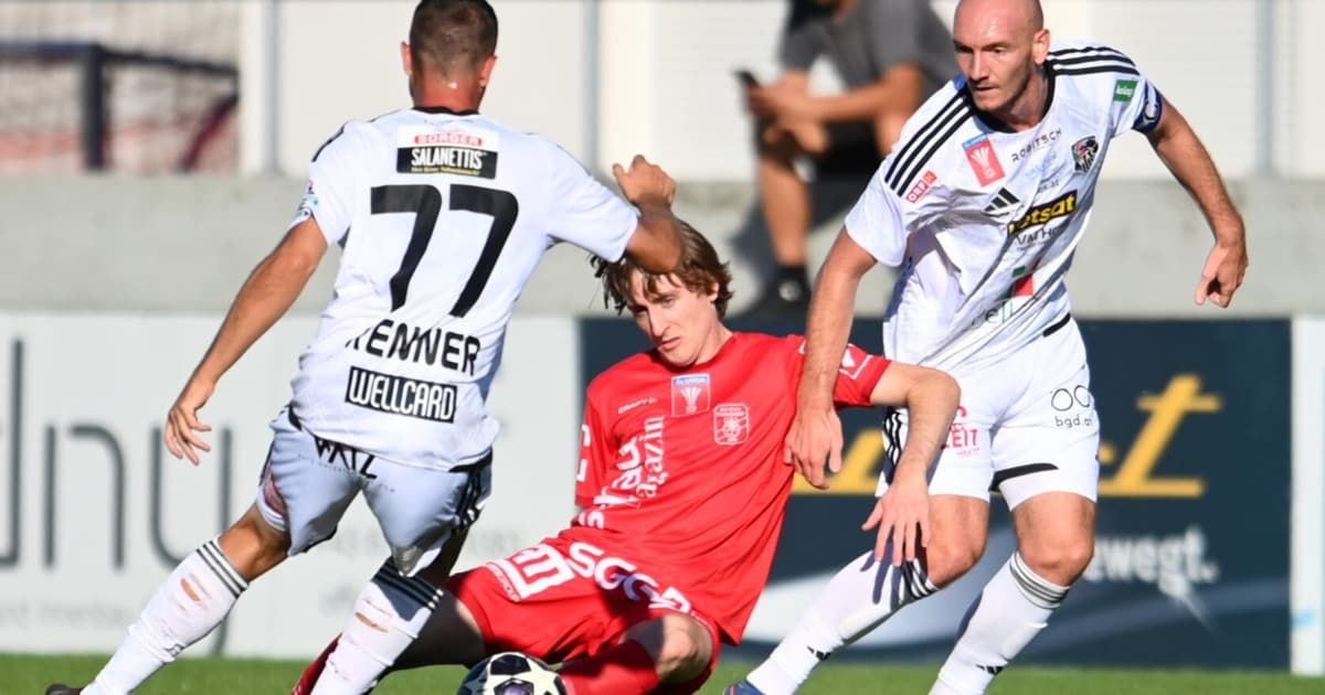 Drei Fußballspieler sind auf dem Spielfeld. Der Spieler in Rot versucht, den Ball zu schießen, während zwei Spieler in Weiß versuchen, ihn zu blockieren.
