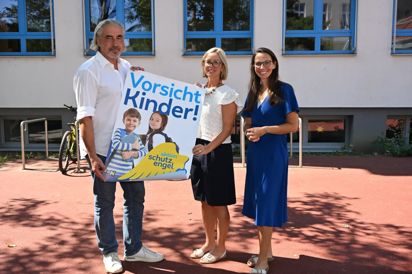 Three adults stand outside a building, holding a sign that reads 'Vorsicht Kinder!' with images of two children. The man wears a white shirt and jeans, while the women wear white and blue dresses.