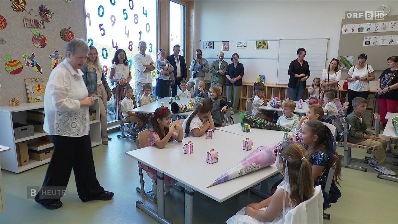 Ein Klassenzimmer mit Kindern an Tischen, Erwachsenen im Hintergrund und einer Wand mit Zahlen und Bildern.