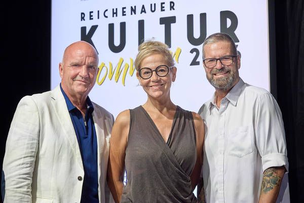 Three individuals stand together in front of a backdrop with the text 'Reichenauer Kultur 2021'. The man on the left wears a white blazer, the woman in the middle wears a sleeveless top, and the man on the right has a tattoo on his arm.