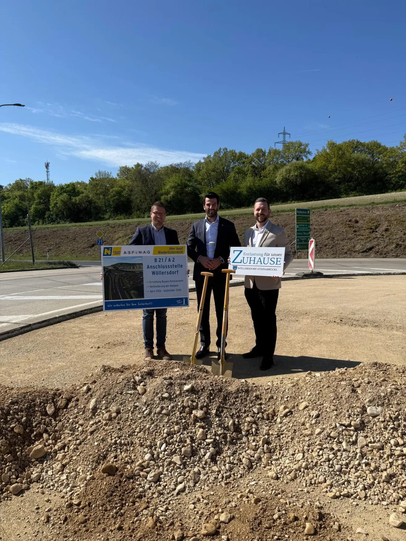 Three men stand on a construction site, holding signs and shovels. The middle man holds a shovel, while the others hold signs. Behind them is a road under construction.