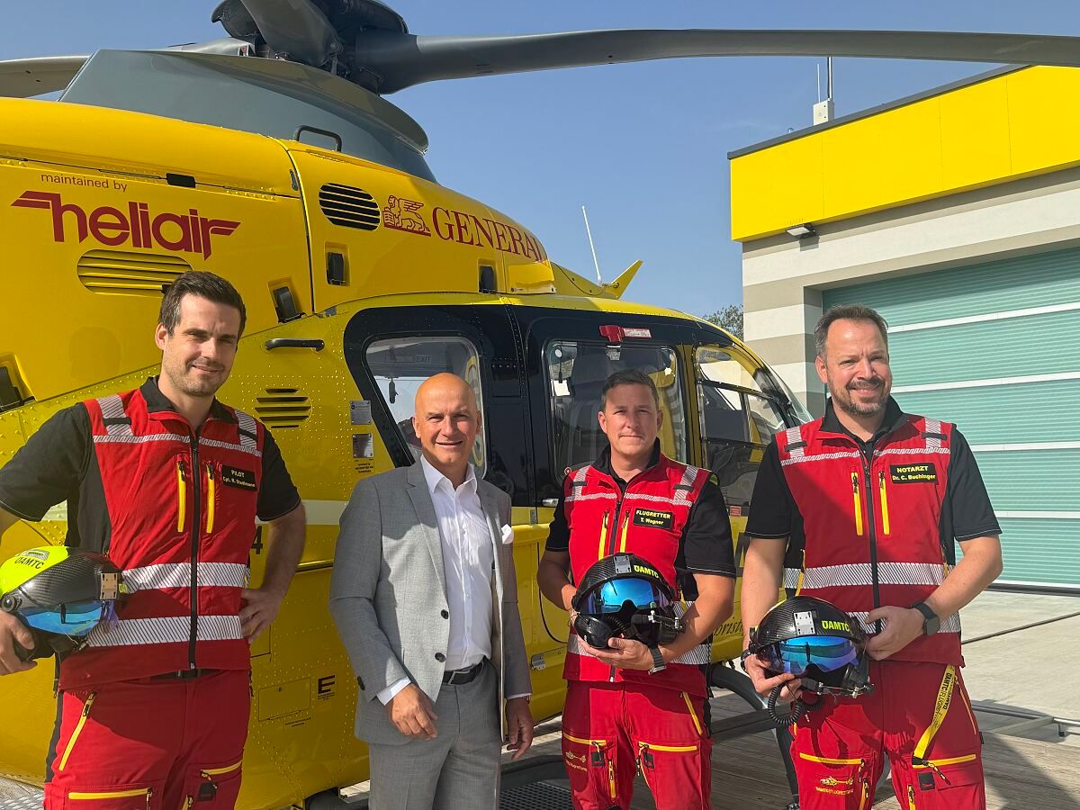 Vier Männer stehen vor einem gelben Hubschrauber. Zwei Männer in roten und weißen Westen halten Helme, und die anderen zwei tragen graue Anzüge. Der Hubschrauber hat das Wort 'General' an seiner Seite.