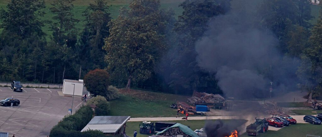 A fire burns in an outdoor area with trees, a building, and parked vehicles. Smoke rises from the fire, and people are seen nearby.