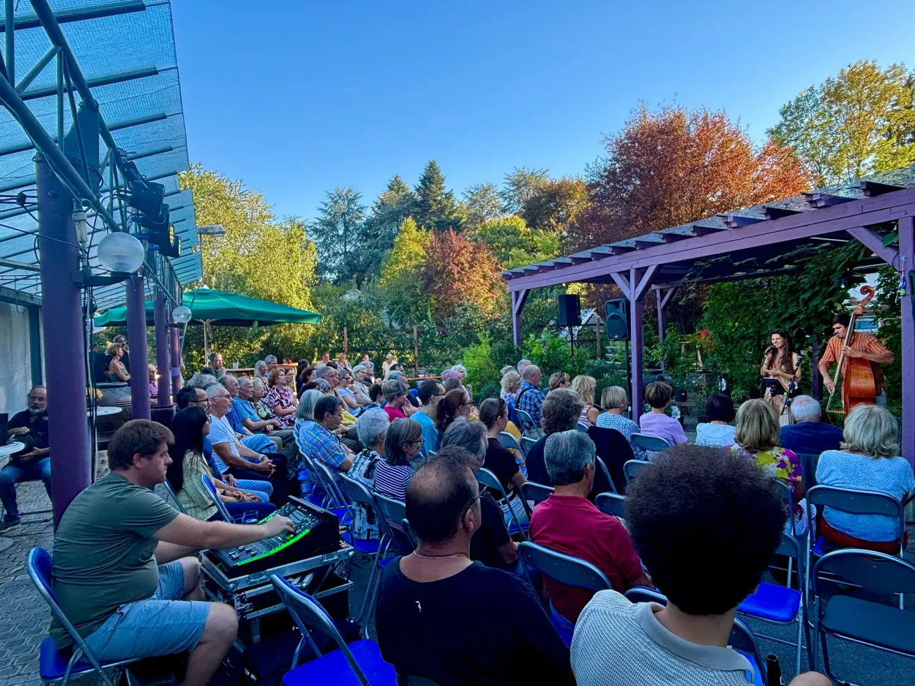 A crowd of people sits in chairs under a purple gazebo, listening to a woman on stage. The setting is outdoors with trees and a clear sky.