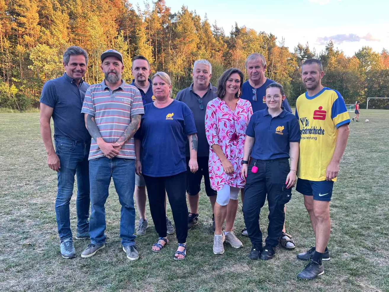 Eine Gruppe von Menschen steht zusammen auf einem Rasen, mit Bäumen im Hintergrund. Sie lächeln und scheinen für ein Foto zu posieren.