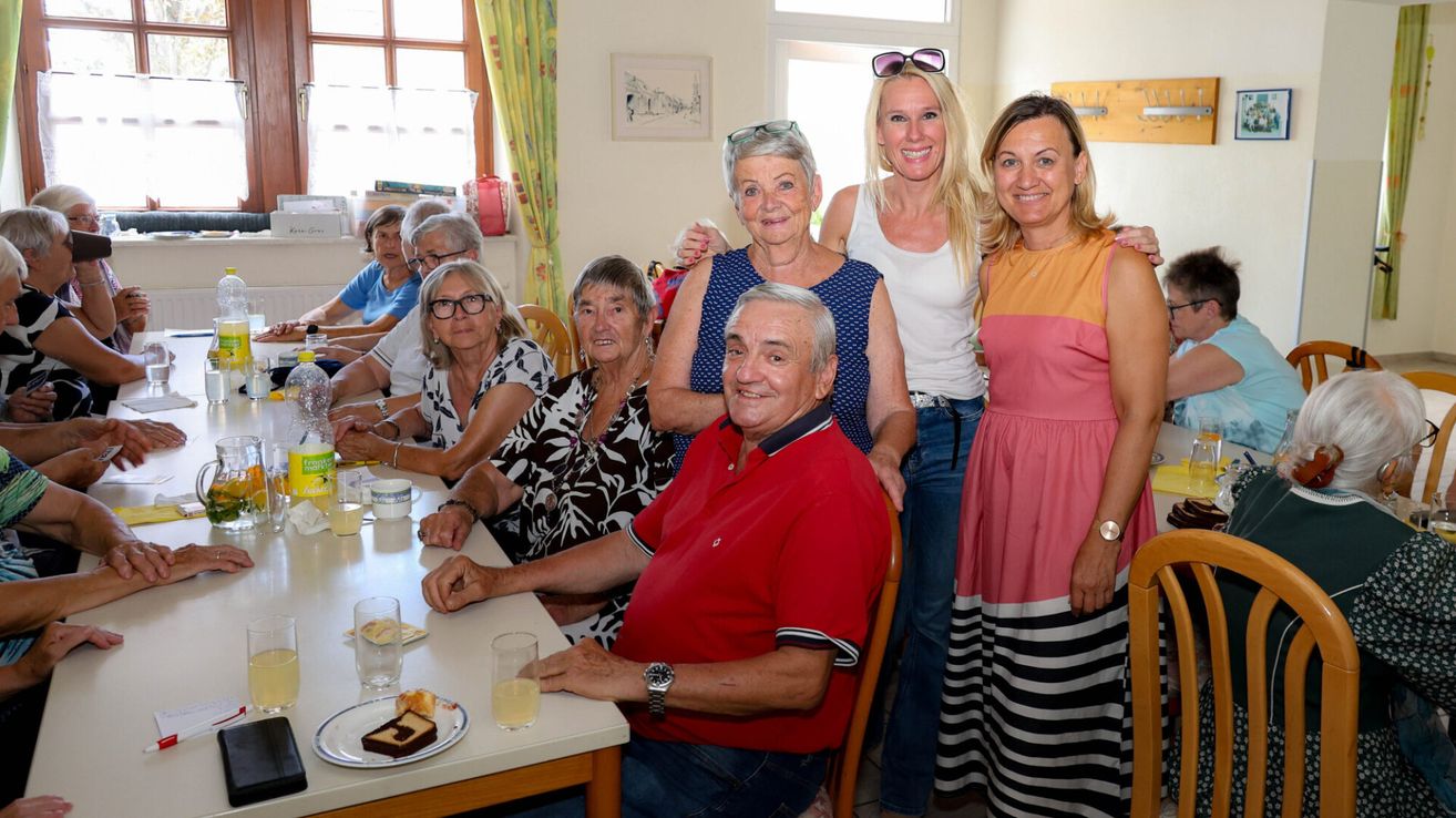 Eine Gruppe älterer Menschen sitzt an einem Tisch und lächelt für ein Foto. Ein Mann trägt ein rotes Hemd und eine Frau in einem blauen Kleid steht hinter ihm.