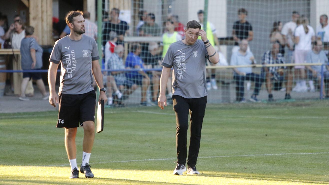 Zwei Männer in grauen T-Shirts gehen auf einem Fußballfeld, einer hält ein Klemmbrett. Ein Mann im Hintergrund berührt seine Stirn. Zuschauer beobachten hinter einem Zaun.