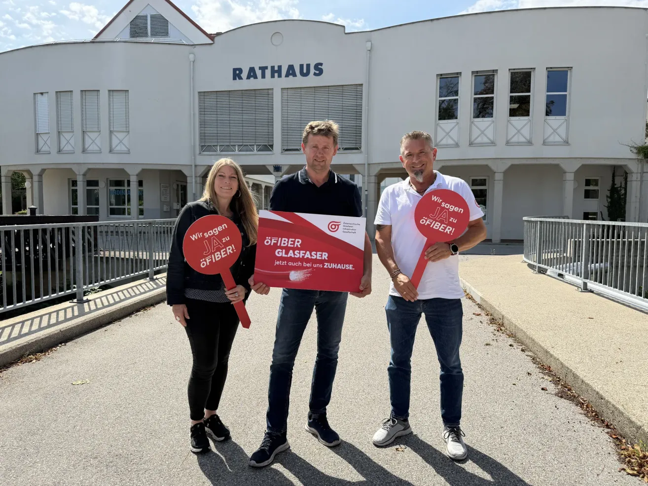 Drei Personen stehen vor einem Gebäude mit dem Wort Rathaus. Sie halten rote Schilder mit der Aufschrift Ja zu Glasfaser.