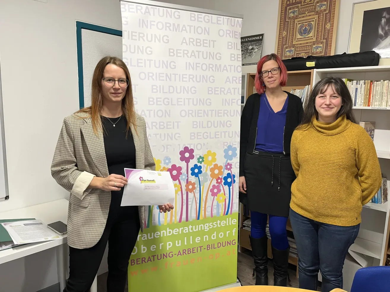 Drei Frauen stehen vor einem Banner, eine hält ein Zertifikat. Das Banner trägt den Text 'Karriereberatung Oberpullendorf'. Dahinter steht ein Bücherregal mit Büchern und ein dekorativer Wandbehang.