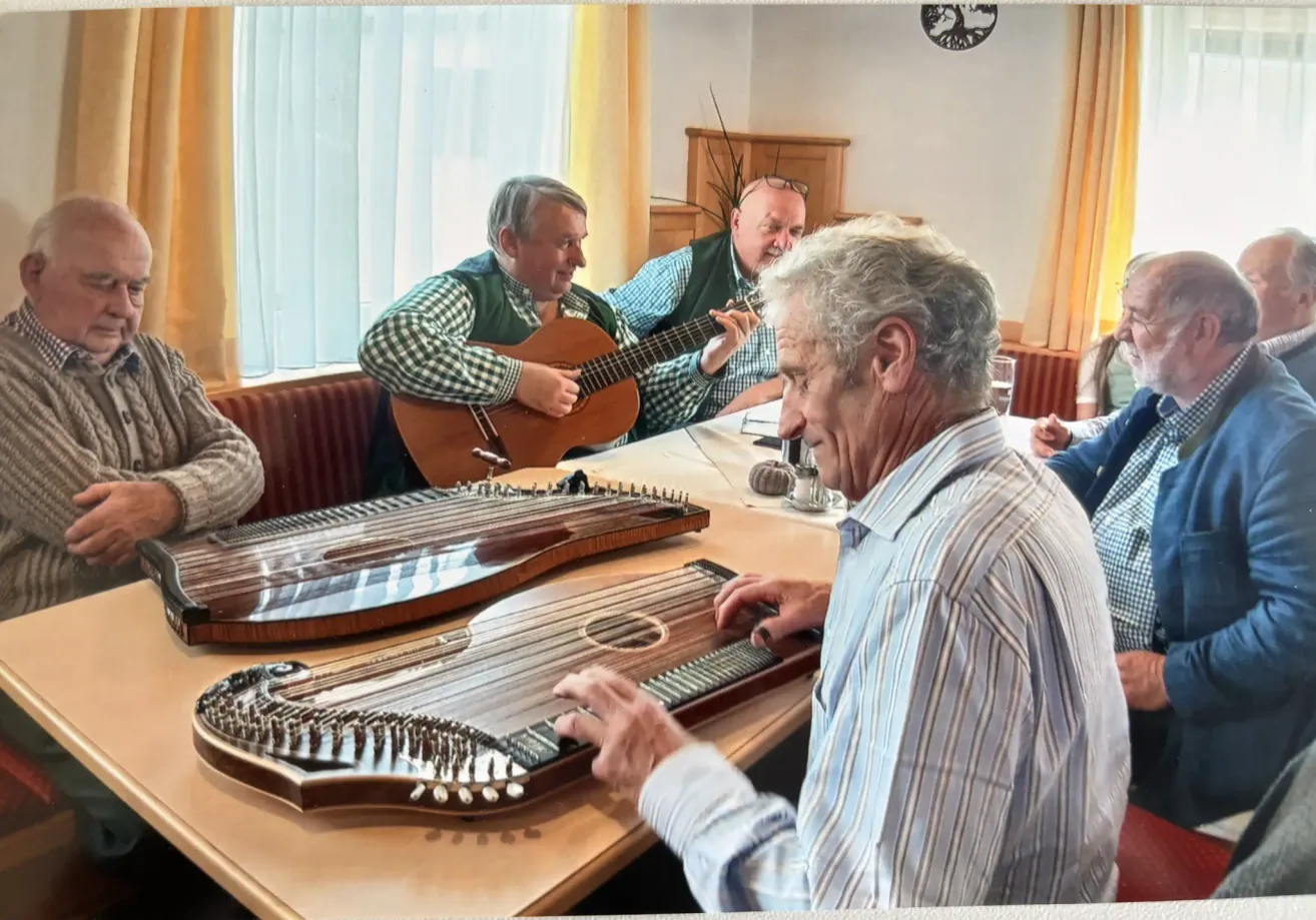 Vier Männer sitzen um einen Tisch herum und spielen Saiteninstrumente. Ein Mann spielt Gitarre, während ein anderer eine Zither spielt. Der Mann vorne lächelt und schaut auf die Zither.