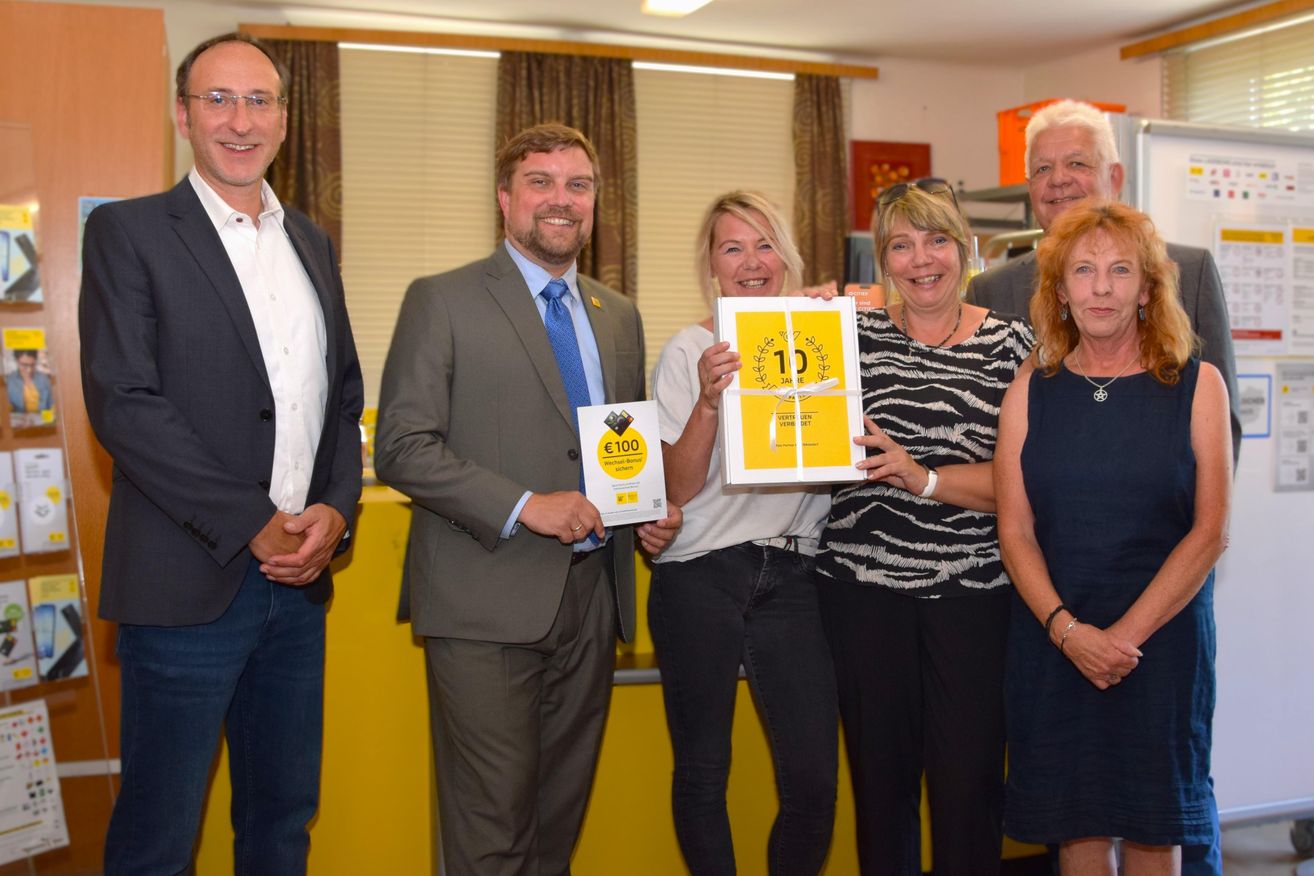 Eine Gruppe von Menschen steht zusammen und lächelt für ein Foto. Ein Mann in Anzug hält ein Zertifikat und eine Frau hält eine Geschenkbox.