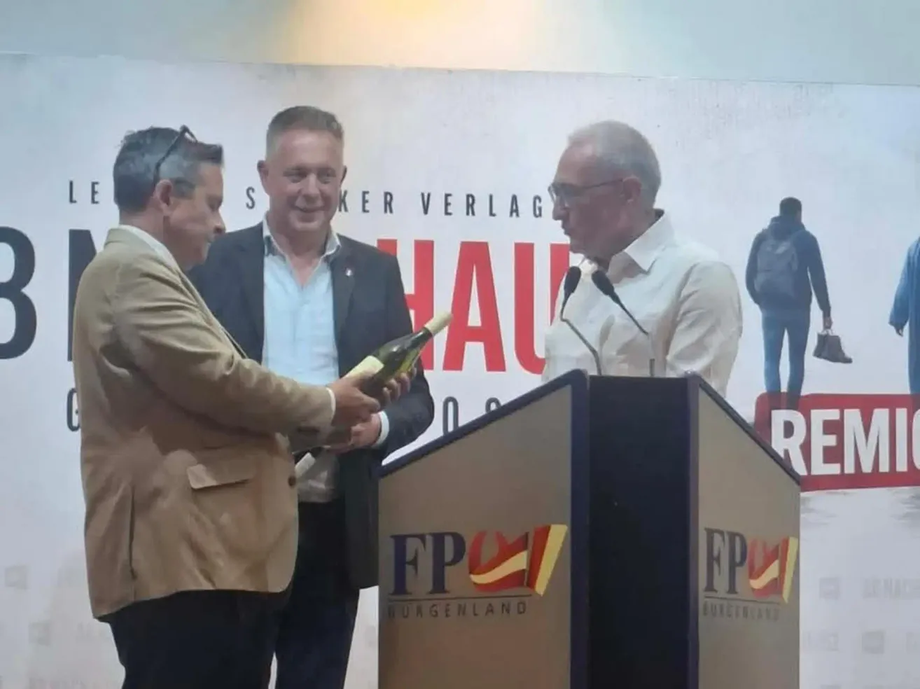 Three men at a podium, one holding a bottle of wine, another smiling, and the third speaking into a microphone. Behind them, a banner reads 'HAUER VERLAG'.