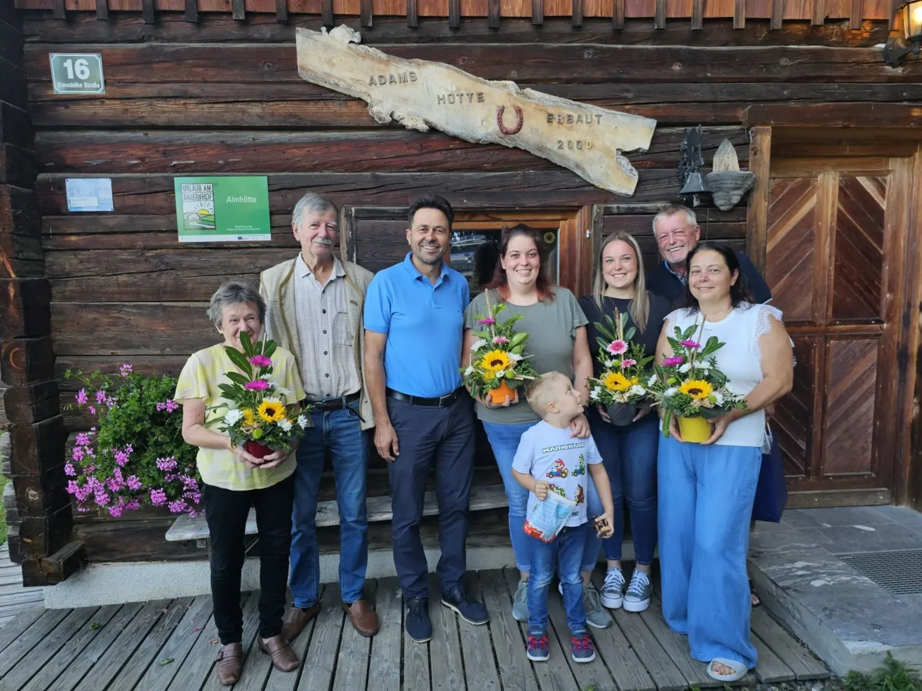Eine Familie steht vor einem Holzchalet, lächelt und hält Blumensträuße. Ein Schild mit dem Text 'ADAMS HUTTE' ist an der Wand hinter ihnen angebracht.
