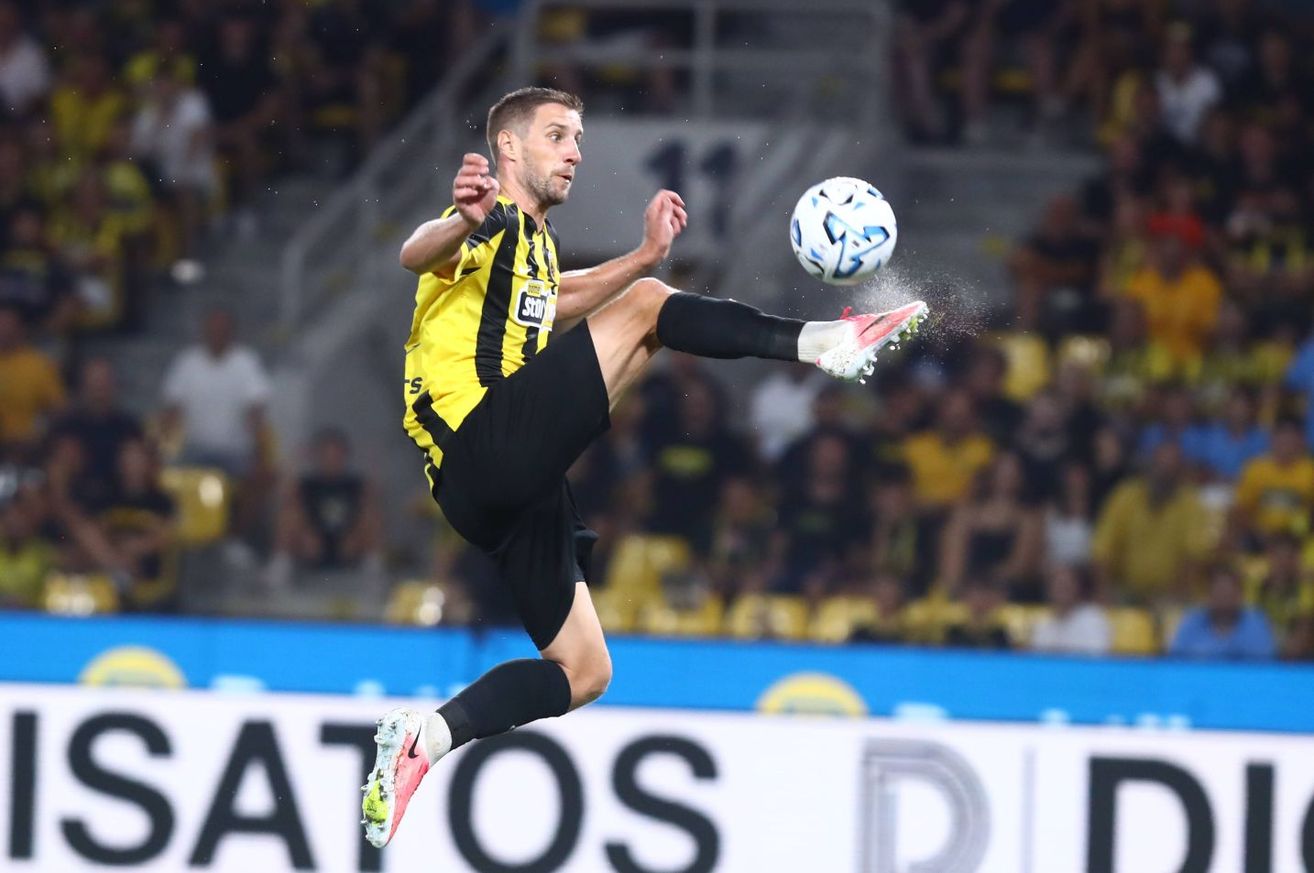 A soccer player in a yellow and black uniform is in mid-air, kicking a soccer ball during a match. The player is wearing Nike cleats.