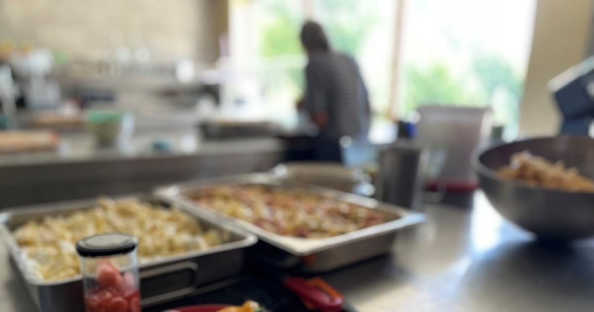 Ein verschwommenes Bild einer Küche mit einer Person, die in der Nähe eines Fensters steht. Zwei Tabletts mit Essen stehen auf dem Tresen.