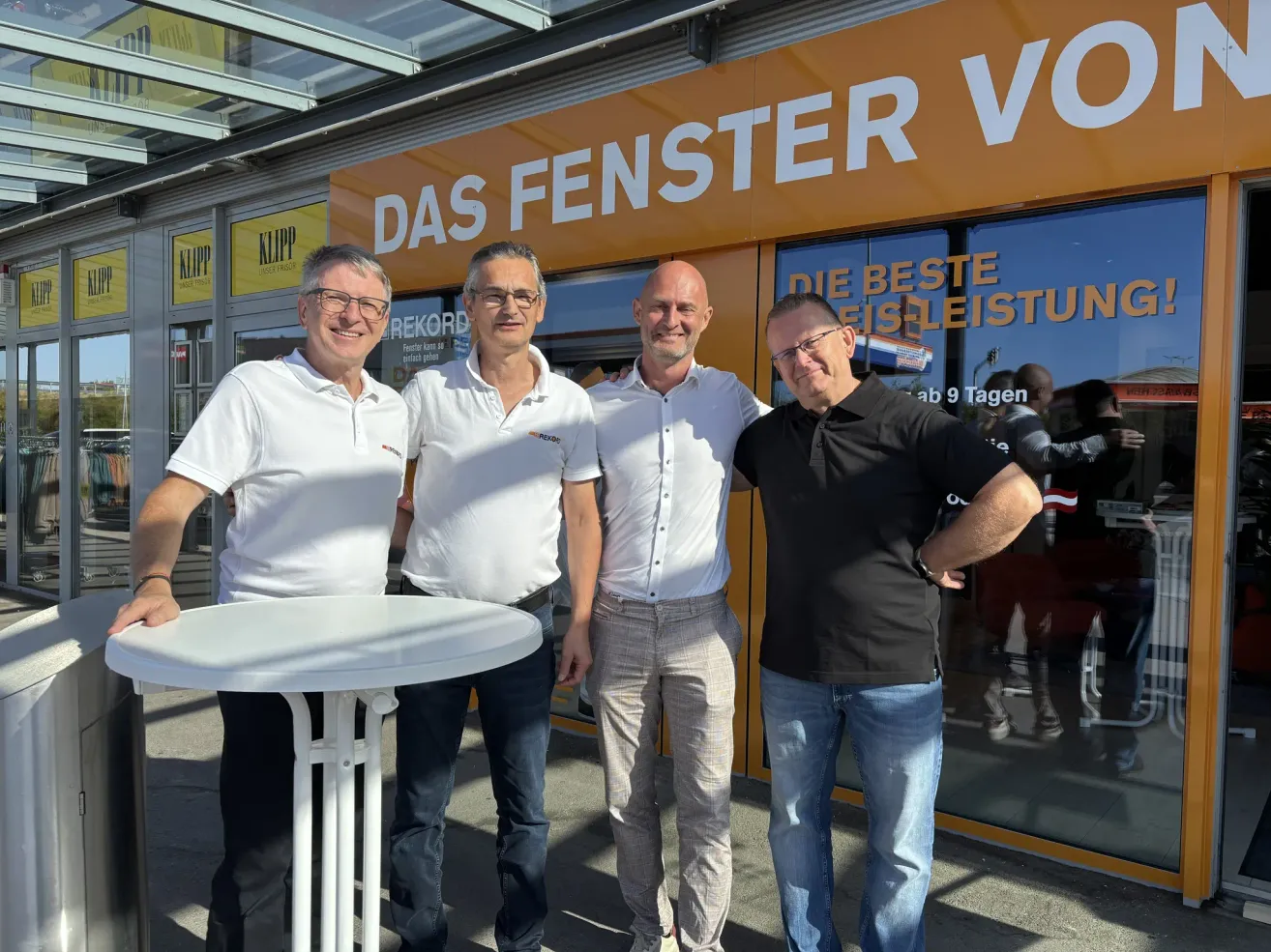 Four men are standing in front of a store with a sign that reads 'DAS FENSTER VO'. They are smiling and appear to be posing for a photo.