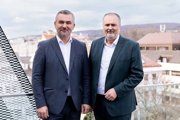 Two men in suits stand on a rooftop balcony, smiling and looking at the camera. Behind them is a cityscape with buildings and a mountain range.