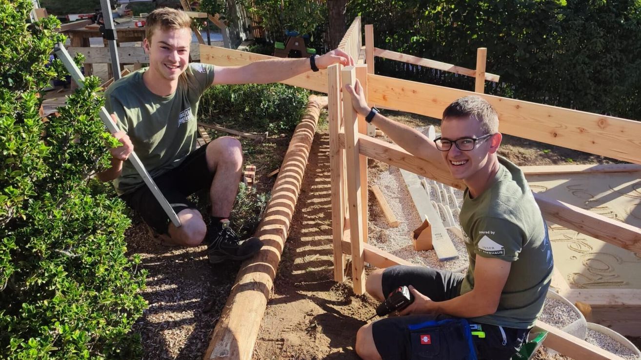 Two men in green shirts are working on a wooden fence in a garden. One is holding a drill, and the other is holding a wooden plank. They are smiling and appear to be enjoying the work.