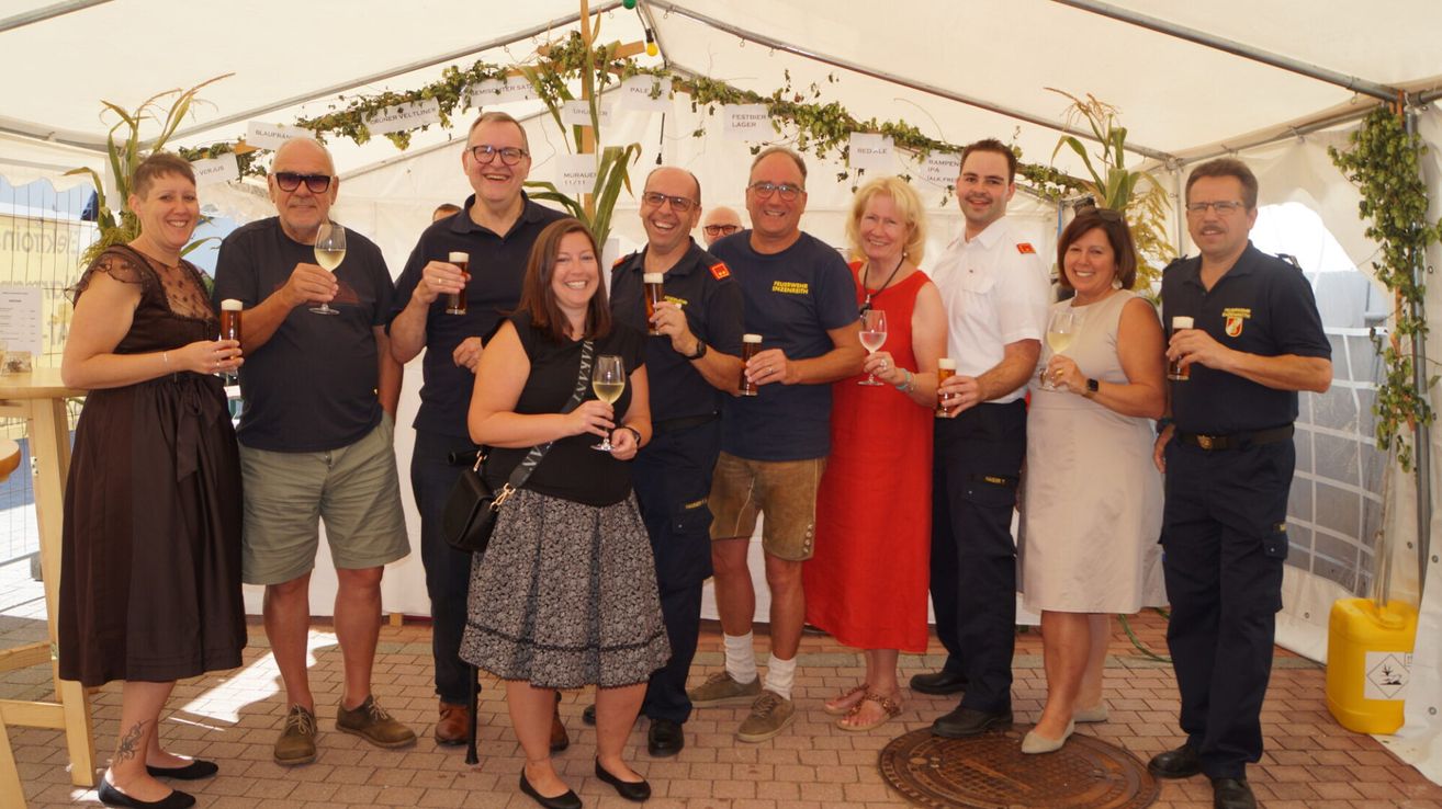 A group of people, possibly at a festival, are holding drinks and smiling for a photo. They are standing in a row under a tent with decorative plants.