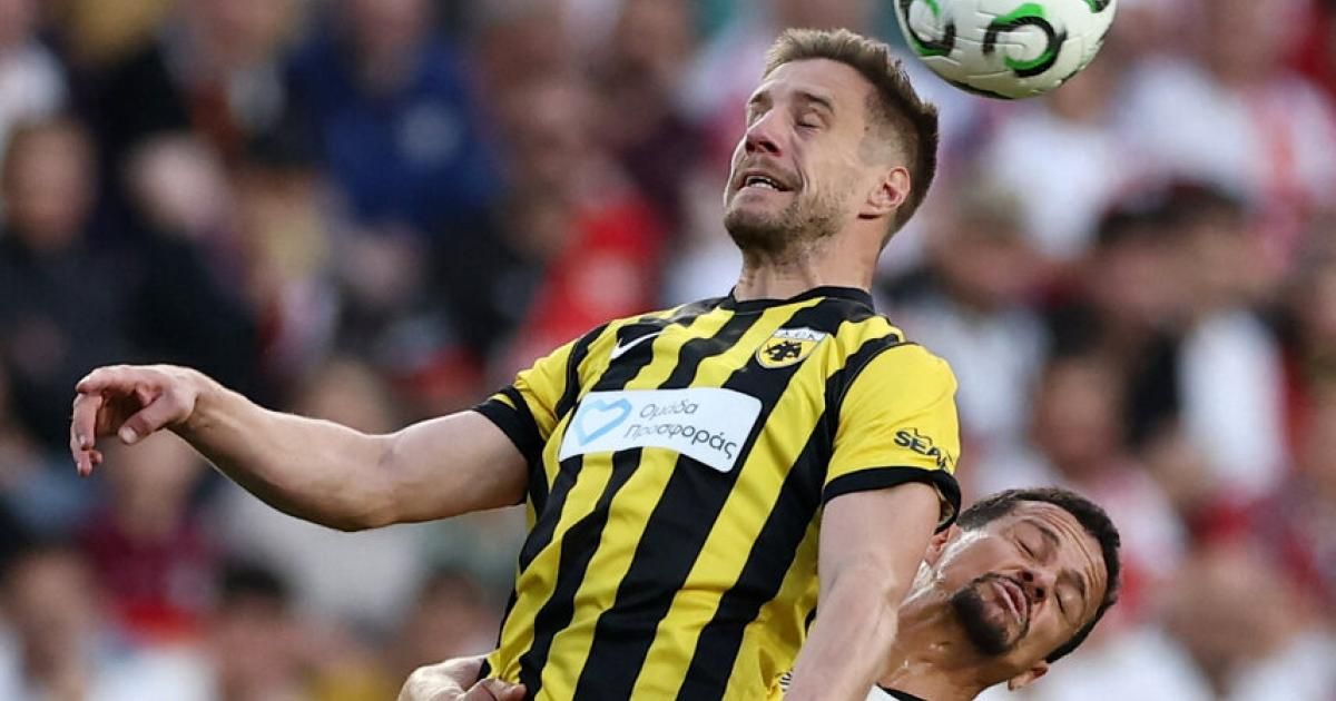 A soccer player in a yellow and black striped jersey is heading a ball while another player looks on.