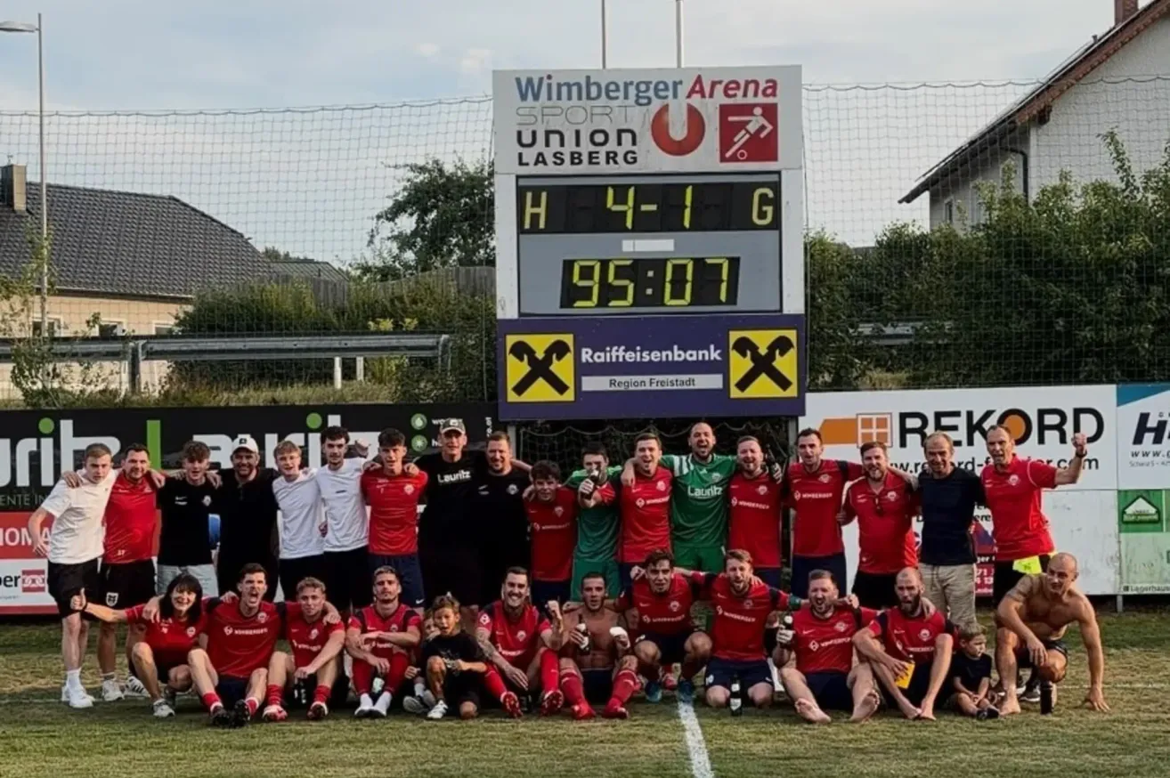 Ein Fußballteam feiert vor einer Anzeigetafel, die einen 4:1-Sieg zeigt. Das Team trägt rote und grüne Trikots, einige Mitglieder tragen Schwarz. Im Hintergrund befindet sich eine Sportarena mit einem Sponsorenlogo.