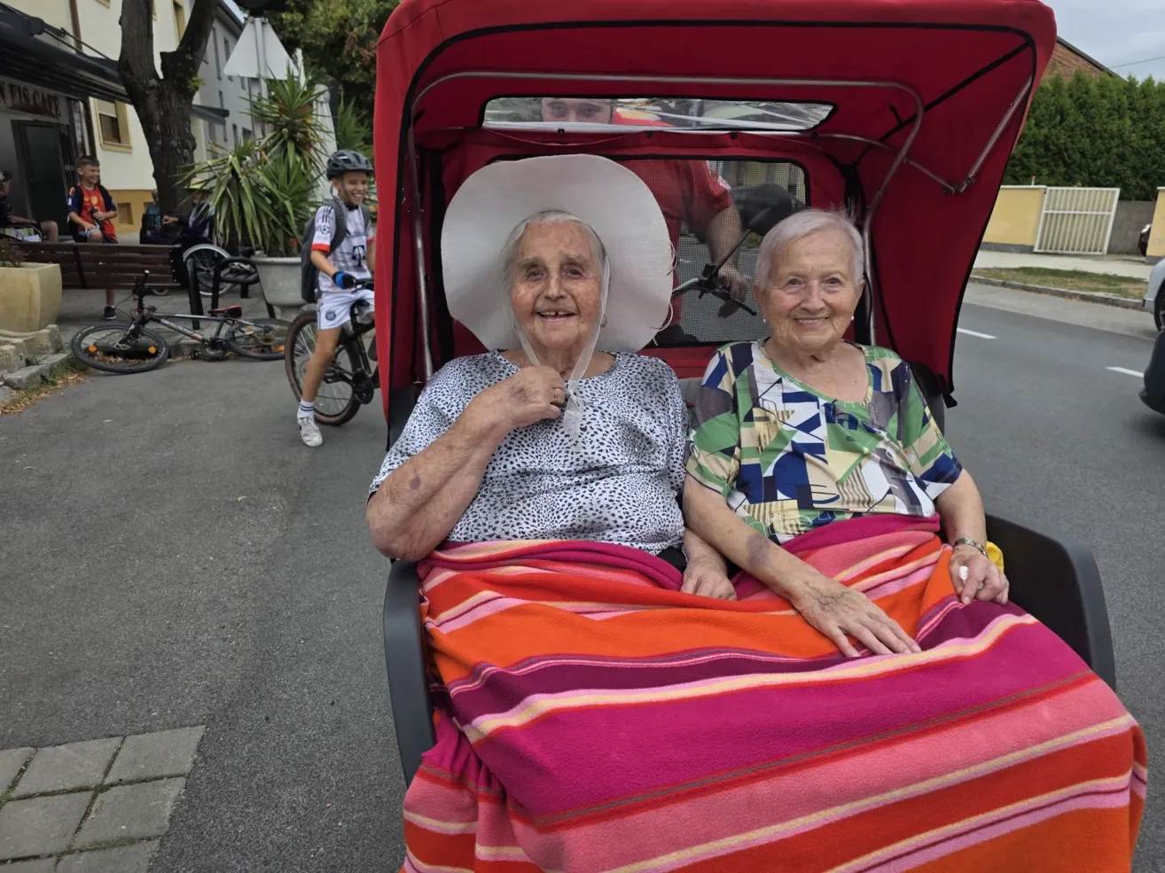 Two elderly women sit in a red tricycle, smiling and covered with a colorful blanket. A child rides a bike in the background.