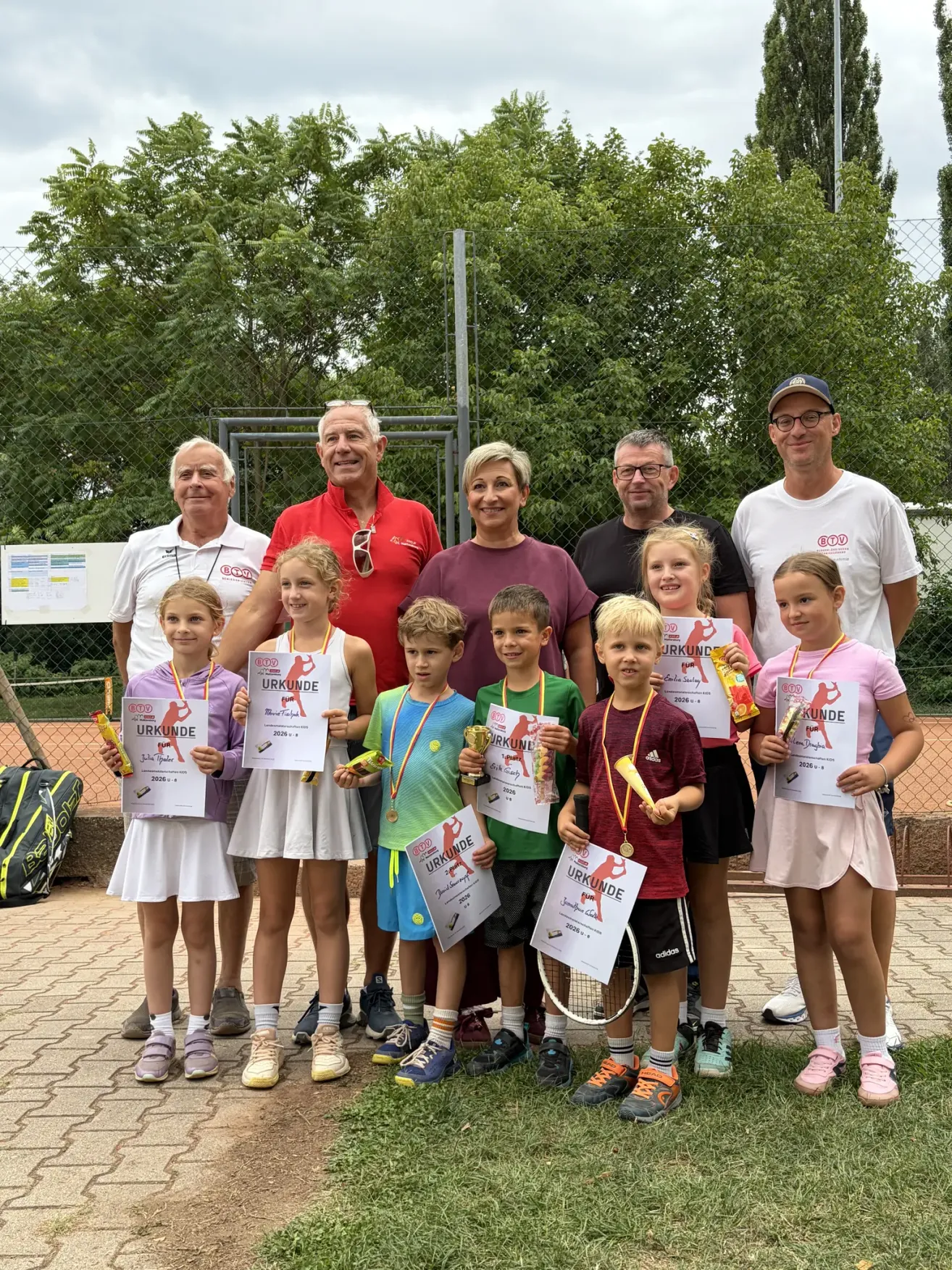A group of people, including adults and children, are standing together on a tennis court, holding certificates and medals. They are all smiling and posing for a photo.