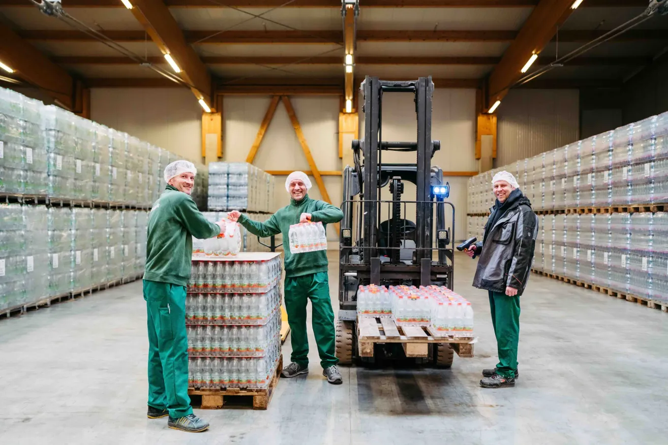Drei Arbeiter in einem Lager, einer hält eine Flasche, ein anderer einen Gabelstapler und ein Dritter lächelt.